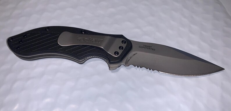 May include: A black and silver folding knife with a partially serrated blade. The knife has a textured black handle and a silver pocket clip. The blade has text that reads "TRIDENT" and "PATENTED".