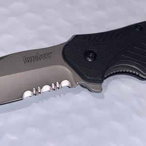 May include: A black and silver folding knife with a partially serrated blade. The blade has a curved edge and the handle is textured for grip. The word "Kershaw" is printed on the blade.