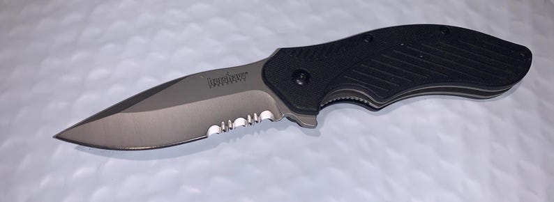 May include: A black and silver folding knife with a partially serrated blade. The blade has a curved edge and the handle is textured for grip. The word "Kershaw" is printed on the blade.