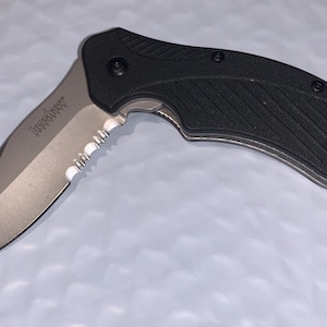 May include: A black and silver folding knife with a partially serrated blade. The knife has a textured black handle and the brand name "Kershaw" is visible on the blade. The blade is approximately 3 inches long.