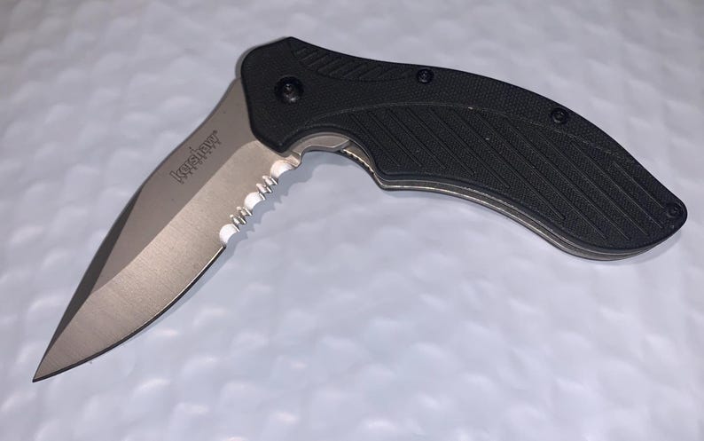 May include: A black and silver folding knife with a partially serrated blade. The knife has a textured black handle and the brand name "Kershaw" is visible on the blade. The blade is approximately 3 inches long.