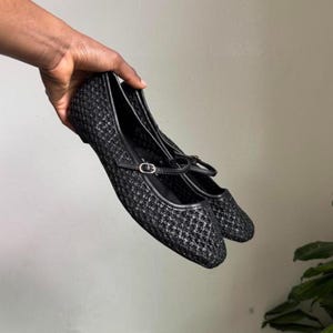 May include: A pair of black mesh Mary Jane flats with a small strap and buckle closure. The shoes have a rounded toe and a thin black trim around the edges. The shoes are held up against a neutral background.