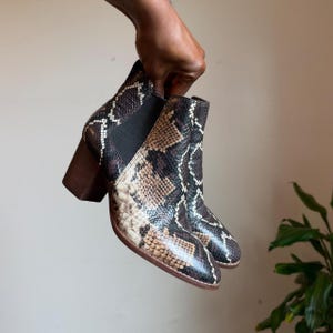 May include: Ankle boots featuring a brown, black, and cream snakeskin pattern. The boots have a rounded toe, a dark brown block heel, and black elastic side panels. The design includes a pull tab at the back of the ankle for easy wear.