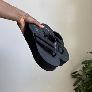 May include: Black platform flip-flops with thick soles and padded straps. The sandals are held up against a neutral background. The flip-flops have a simple, minimalist design, suitable for casual wear.