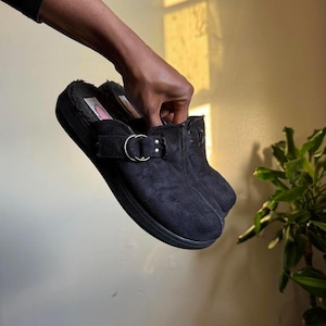 May include: Black suede-effect clogs with a faux fur lining. The clogs feature a buckle detail with a silver-coloured metal ring. The soles are black and the shoes appear unworn.