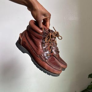 May include: A pair of brown leather ankle boots featuring a padded collar, a moccasin-style stitched toe, and dark brown laces. The boots have a thick, dark rubber lug sole and a small embossed logo on the side of the heel area of the leather upper.