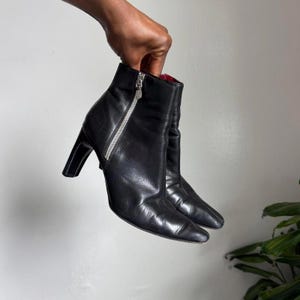 以下が含まれることがあります： 黒い革のアンクルブーツのペア。つま先が尖っており、高いブロックヒールが付いています。ブーツは、側面にシルバーのジッパー開閉部と赤い内側を備えています。ブーツは手で持たれています。