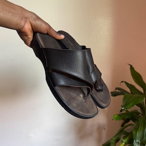 May include: A pair of dark brown leather slide sandals featuring a wide, overlapping strap design across the top and a contoured footbed. The sandals have a thick, solid black sole and a toe post construction, providing a simple and functional design.