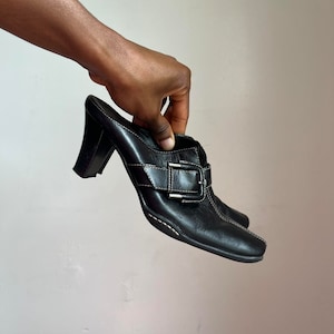 May include: Black leather heeled mules with a buckle detail. The heels are about 3 inches tall.