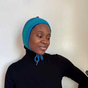 Puede incluir: Un gorro de ganchillo hecho a mano en tonos turquesa y azul, con un pequeño acento magenta en la parte superior. El gorro tiene lazos debajo de la barbilla. El artículo es probablemente un accesorio de moda o parte de un disfraz.