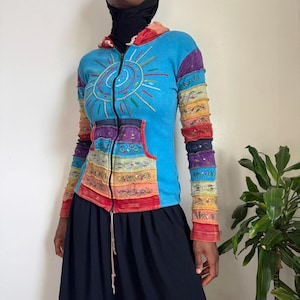May include: A blue zip-up hoodie with a sun design embroidered on the front. The sleeves and pocket feature colourful patchwork. The hoodie has a drawstring and a black zip. The garment is worn with a black skirt.