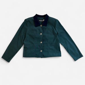 May include: A dark teal jacket featuring a contrasting navy blue collar, long sleeves, and a front button closure with four metallic buttons. The garment includes horizontal and vertical stitched panel detailing across the front bodice.