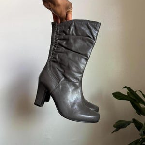 May include: Gray leather mid-calf boots with a block heel. The boots have a ruched design on the side and a rounded toe. The heel height is approximately 3 inches.
