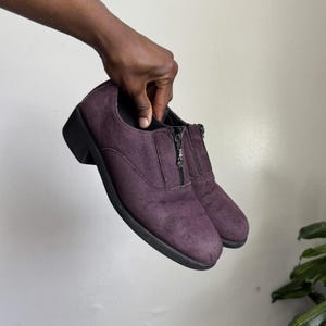 May include: A pair of plum-colored shoes with a zipper closure. The shoes have a low heel and a rounded toe. The material appears to be suede or a similar textured fabric. The soles are black.