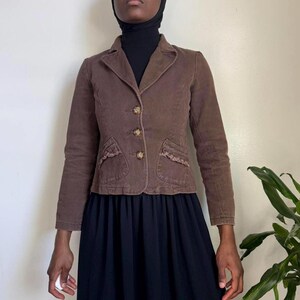 May include: Brown corduroy blazer with three gold-colored buttons and two front pockets with decorative trim. The blazer has a classic collar and long sleeves. The garment is worn over a black top and a dark blue skirt.