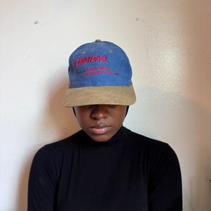 May include: A blue baseball cap with a tan brim featuring the red embroidered text COMPAQ and the smaller red text Innovation that speaks for itself. The cap has a structured crown and a curved visor, with small ventilation eyelets on the top panels.