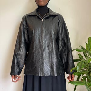 Puede incluir: Una chaqueta de cuero negro con un diseño de patchwork con costuras visibles, cuello en punta y un cierre de cremallera metálica plateada de arriba a abajo en la parte delantera. La chaqueta tiene mangas largas y un dobladillo recto.