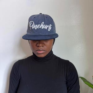 May include: A denim blue flat-brimmed baseball cap with the word "Pinehurst" embroidered in white script across the front. The cap has a structured crown and a dark blue brim. The person is wearing a black turtleneck.