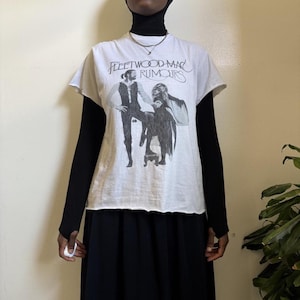 May include: A white short-sleeved t-shirt featuring a black graphic print of the Fleetwood Mac Rumours album cover. The shirt has a raw, unfinished hemline at the sleeves and waist. It is layered over a black long-sleeved top and a black skirt.