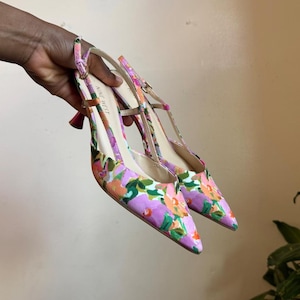 May include: A pair of Nine West pointed-toe slingback heels featuring a vibrant floral pattern in shades of purple, orange, and green. The shoes have a thin adjustable ankle strap with a gold-tone buckle and a slender heel, with the brand name printed.