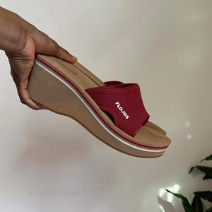 May include: A red wedge sandal with the brand name "FLOJOS" on the strap. The sandal features a tan sole with white and red trim. The wedge heel offers lift and support.