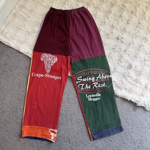 May include: Colour-blocked lounge trousers featuring a maroon waistband, a red left leg with a white Grape-Stomper graphic, and a green right leg with white Louisville Slugger text that reads Swing Above The Rest, finished with orange and blue accents.