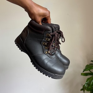 May include: A pair of dark brown ankle-height boots featuring a smooth synthetic exterior, a padded collar, and dark brown laces threaded through metallic eyelets. The boots have a thick, textured rubber sole and a rounded toe design.