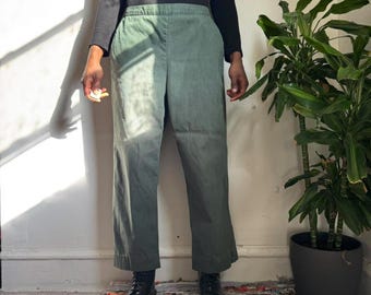 May include: Sage green pants with a wide leg design. The pants have a high waist and are made of a soft, textured fabric. The pants are paired with black boots and a black long-sleeve shirt. A houseplant is in the background.