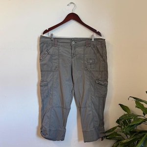 May include: Gray cargo capri pants featuring a button and zipper fly closure, multiple utility pockets on the front and sides, belt loops, and adjustable hem ties. The pants are displayed on a dark brown plastic hanger against a plain wall.