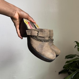 May include: A pair of brown clogs with a platform sole. The clogs feature a buckle strap and a decorative chain detail. The shoes have a distressed, worn appearance, and the heel is approximately 3 inches high.