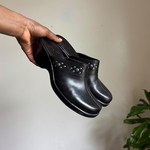 May include: A pair of black slip-on wedge mules featuring a rounded toe, a thick sole, and a decorative band across the upper vamp adorned with small, circular silver-toned metal studs arranged in a repeating pattern along the leather-like surface.