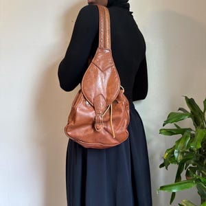 May include: A medium-sized, cognac-colored leather sling backpack featuring a teardrop-shaped front flap with a brass-toned buckle closure. The bag has a single vertical shoulder strap, a zippered main compartment, and visible stitching details.
