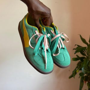 May include: A pair of green suede sneakers featuring a yellow leather side stripe, light pink laces, and a dark brown rubber sole. The Puma brand logo is visible on the tongue of the shoe. The sneakers have a low-top design with a rounded toe.
