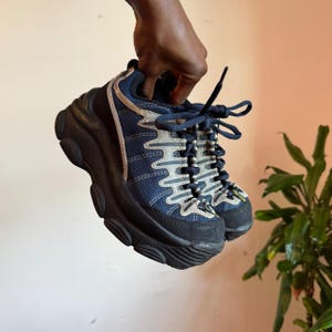 May include: A pair of navy blue and black sneakers with a thick, chunky black rubber sole. The upper features a dark blue mesh material with light gray wavy overlays, navy blue laces, and a small yellow tag near the toe area of the shoe.