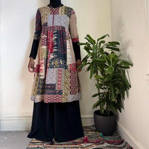 Puede incluir: Una túnica patchwork con mangas largas, con una mezcla de patrones en rojo, negro, beige y azul marino. El vestido se combina con pantalones negros de pernera ancha y un cuello alto negro. Una planta en una maceta oscura está en el fondo.