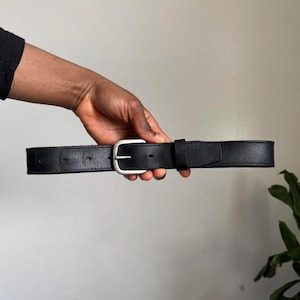May include: A black leather belt with a silver-colored buckle. The belt is held horizontally, showcasing its simple design and construction. The buckle is a classic, rectangular shape.