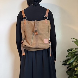 May include: A tan canvas backpack featuring brown leather-look straps, silver-toned metal rings, and a vertical central strap with a buckle. The bag includes a zipped pocket on the front, a small rectangular patch, and adjustable side straps with metal hardware.