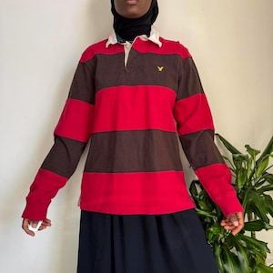 May include: A red and brown striped long-sleeved rugby shirt with a white collar and a small yellow bird logo on the chest.