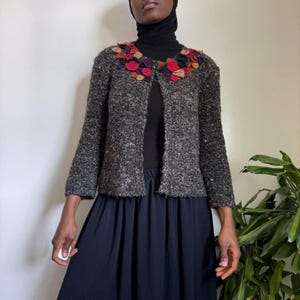 May include: A gray tweed jacket with a floral collar featuring red, purple, and orange fabric flowers. The jacket has long sleeves and a buttonless front. The model is wearing a black top and a long navy blue skirt.