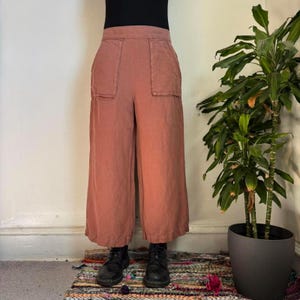 May include: Dusty rose-colored wide-leg pants with two front pockets. The pants are made of a soft fabric and have a high waist. The model is wearing black boots. The pants are part of a casual clothing collection.