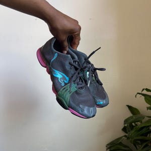 May include: A pair of dark grey athletic trainers featuring a bright blue Puma logo on the side, a green midfoot support cage, and a vibrant pink sole. The shoes have dark grey laces and a mesh upper construction with a low-top design profile.
