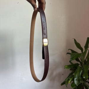 Puede incluir: Un cinturón marrón oscuro con un patrón texturizado de piel de lagarto y un reverso liso de color beige. El cinturón presenta una hebilla rectangular de metal dorado con esquinas redondeadas y acabado pulido, sujeta por una trabilla a juego.