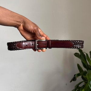 May include: A dark brown leather belt with a woven design and a silver-colored buckle. The belt is held up against a neutral background, showcasing its texture and craftsmanship. A classic accessory.