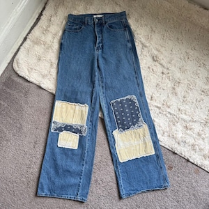 Könnte beinhalten: Hoch taillierte blaue Jeans aus Denim mit weitem Bein und ausgefransten Patchwork-Details an den Knien. Die Flicken bestehen aus cremefarbenem und dunkelblauem Stoff mit Bandana-Mustern und sind auf die Vorderseite der Hosenbeine genäht.