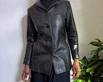 Könnte beinhalten: Ein schwarzer Lederblazer mit klassischem Design. Der Blazer hat einen Reverskragen, drei Knöpfe und eine taillierte Passform. Die Jacke wird über einem schwarzen Rollkragenpullover und einem langen schwarzen Rock getragen.