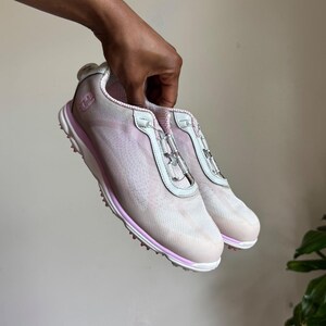 May include: A pair of light pink and white athletic shoes featuring a mesh upper, a white midsole with a pink accent stripe, and a specialized lacing system with clear plastic hardware. The side of the shoe displays a small white logo emblem.