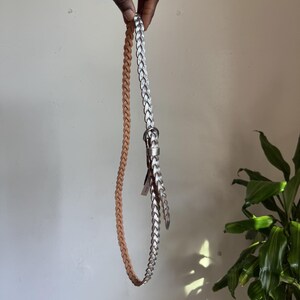 May include: A thin braided belt featuring two distinct colors, with one half in a light tan shade and the other half in a metallic silver finish. The belt includes a small silver-toned metal buckle and a matching silver-toned belt loop keeper.