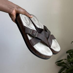 May include: Brown leather sandals with a cork sole and silver buckles. The sandals have a criss-cross strap design and a white footbed. The brand name "Bass" is visible on the footbed. These sandals are a casual footwear option.