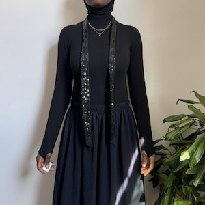 May include: A black turtleneck top and a dark blue skirt. A long, black sequined scarf hangs down the front. A delicate gold necklace is visible. The outfit is accessorized with a black head covering.