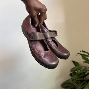 May include: A pair of dark brown leather Mary Jane shoes featuring a textured, crinkled finish, a rounded toe, and a single adjustable strap across the instep with a hook-and-loop closure. The shoes have a low profile and a dark, flat rubber sole.